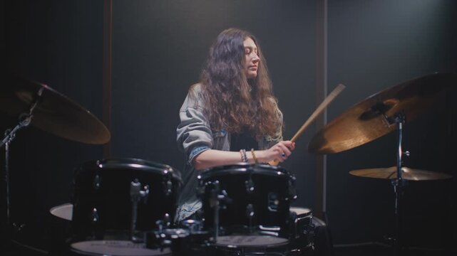 Drummer girl playing drums in music rehearsal studio filled. Rock band music artist learning a drum solo. Young woman with long curly hair, fill or break on drum kit. Female drumming in room