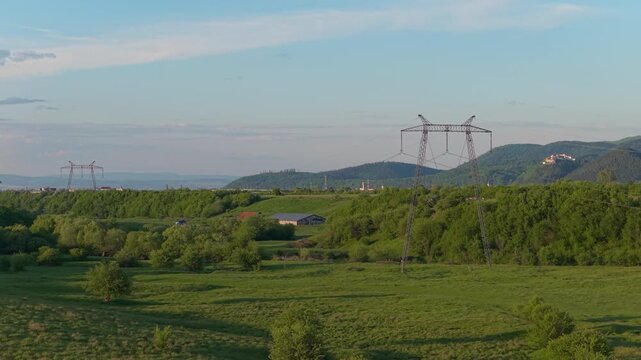 Sweeping high tension power lines over a serene green valley with Rasnov city and citadel in the background, smooth lateral aerial tracking