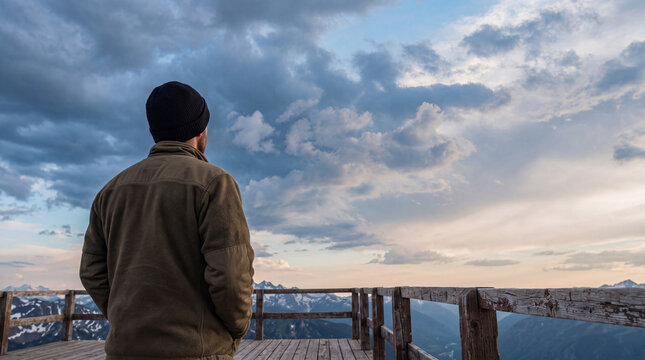 Man on a mountain viewpoint observing the majestic, cloudy sky and expansive snow-capped peaks below.
