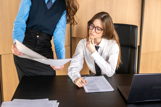 Two businesswomen discussing and reviewing reports and agreements, standing and sitting at a desk with a laptop and documents, symbolizing teamwork and office work