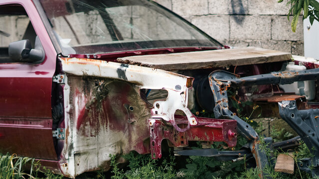 a salvaged red car body sitting in tall grass with its front end missing and a wooden board placed over the engine compartment
