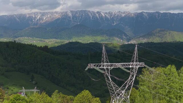 Majestic sunlit electricity pylon in dramatic Moieciu valley with Piatra Craiului mountain peaks in background, slow descending aerial view, graded 4K