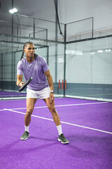 Obraz premium Man playing on purple padel court in purple shirt white shorts holding paddle by glass panels