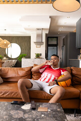 Obraz premium Man sitting on brown sofa in living-room wearing red jersey, holding basketball and sipping glass