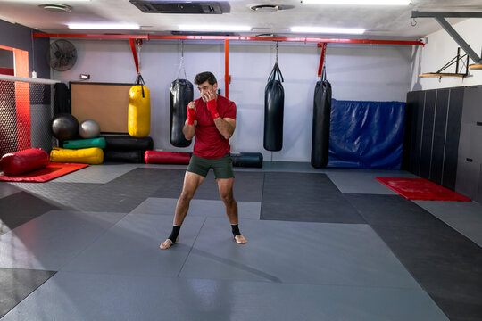Man shadowboxing on padded training floor in martial arts gym near heavy bags wearing red wraps