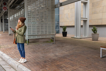 Fototapeta premium Mature woman standing at transit stop near curb, holding phone, wearing beanie and bag, copy space
