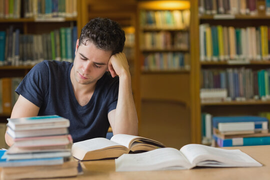 Adult male student sitting at library wooden table reading open textbooks, closed book stack nearby