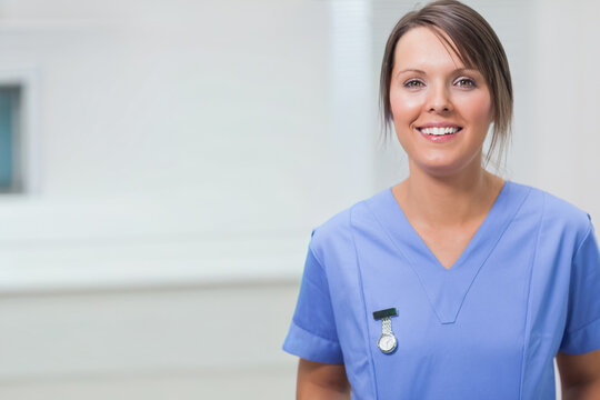 Female nurse standing right in clinic near blinds wearing lightblue scrubtop, fobwatch, copy space