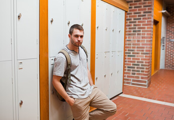 Obraz premium Male student leaning against gray lockers in school corridor, carrying olive backpack, copy space