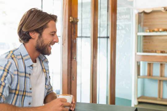 Adult man sitting at cafe counter holding white ceramic cup with latte art, wearing plaid shirt