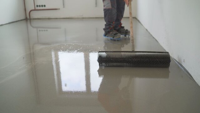 Construction worker applying self leveling screed with spiked roller on concrete floor