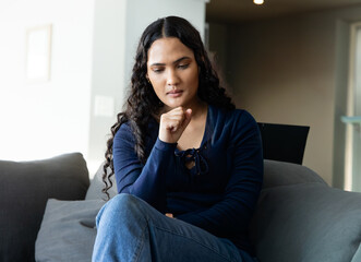 Fototapeta premium Adult woman sitting on gray sofa in living room, wearing navy blouse, holding chin near laptop