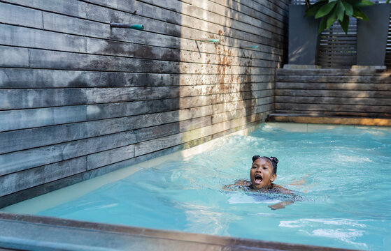 African American child in dark swimsuit swimming in private pool by wooden slat wall, copy space