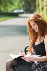 Fototapeta premium Woman sitting on park bench writing with pen in open notebook wearing plaid shirt, copy space