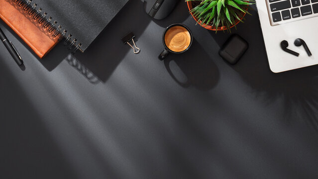 A dark desktop with laptop, black wireless earbuds, leather and spiral notebooks, succulent plant, pen, mouse and espresso coffee cup on dark background with soft lighting. Top view with copy space.