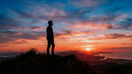Fototapeta premium Man silhouette standing on a mountain peak looking at a beautiful colorful sunset over the ocean and distant hills.