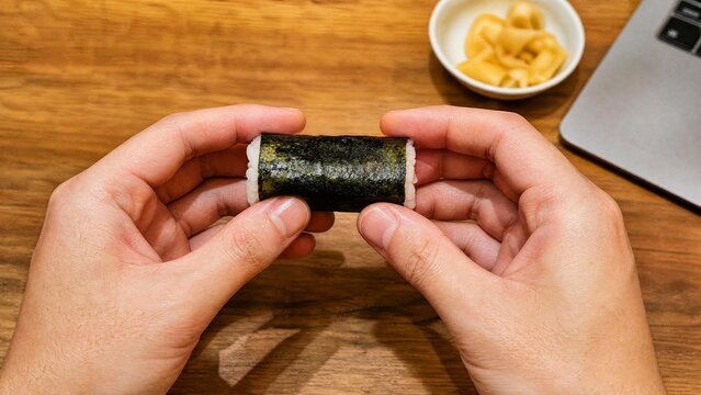 Hands Holding Fresh Sushi Maki Roll at Workspace with Laptop and Ginger