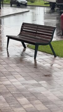 Empty wooden bench on a wet sidewalk during rain in a public outdoor space, quiet urban rest area, city park furniture, rainy day mood.
