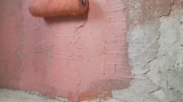 Close up of paint roller applying red orange coat on textured concrete surface. Home improvement or construction site background with cinder blocks. DIY renovation project concept
