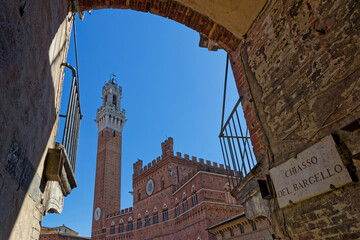 Fototapeta premium SIENA, ITALY, April 25, 2026 : Palazzo Pubblico with its tall Torre del Mangia seen through the frame of an old street arch surrounding Piazza del Campo
