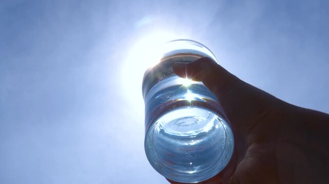 transparent glass glass of water with pure mineral water in hand against the sky with bright glare of the sun