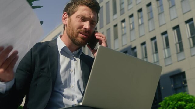 Angry multitasking Caucasian businessman arguing mobile phone city holding documents laptop frustrated male business man employer bad results conflict negotiation work crisis stress outdoors downtown