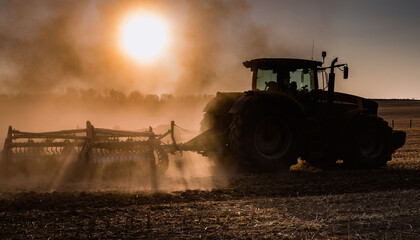Fototapeta premium Powerful tractor silhouetted against a golden sunset working a dusty field, symbolizing modern agriculture and hard work. A dramatic, cinematic shot ideal for farming and industry visuals.