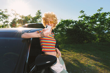 Fototapeta premium Smiling woman in a striped top leans on a car, taking a selfie with a smartphone. Outdoor scene with bright sun, retro film color, and casual fashion mood, under warm golden light and a breezy