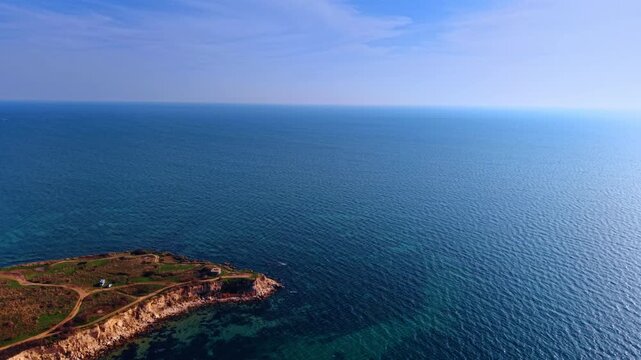 Rocky coastal headland with green fields jutting into calm blue sea. Dirt paths cross a flat green rocky cape ending in dramatic cliffs that drop into clear blue coastal water.