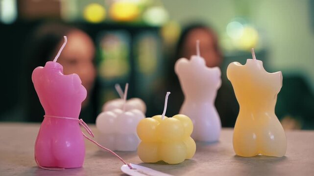 Various colorful shaped handmade candles displayed on a tabletop in a workshop setting with defocused background.