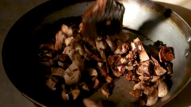 Fresh sliced mushrooms being fried in a hot metal pan with wooden spatula