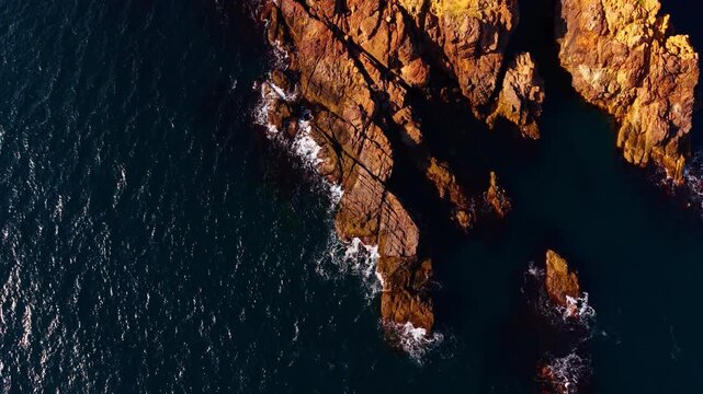 Aerial golden layered red rocks protruding into dark teal sea. Golden sidelight strikes red rock formations as they reach diagonally into dark teal sea with white foam at the edges.