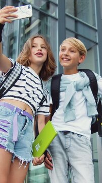 Cute funny small teens boy and girl standing near school building with books and backpacks while doing mocking faces to smartphone camera while taking selfie photos. School concept. Slow motion
