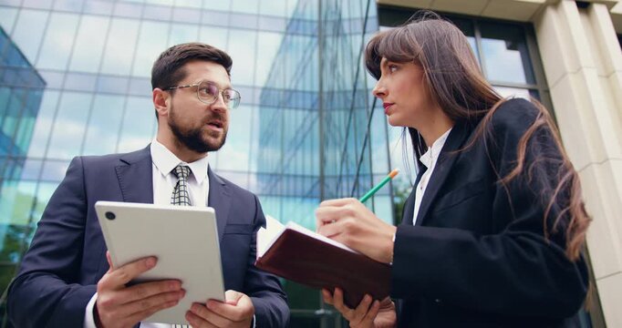 Man and woman working together outdoors, reviewing notes and tablet data in a collaborative business setting