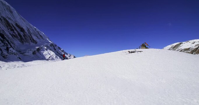 A trekker in a red jacket walks along the snowy shore of stunning Tilicho Lake, framed by towering Himalayan peaks and a brilliant blue sky.