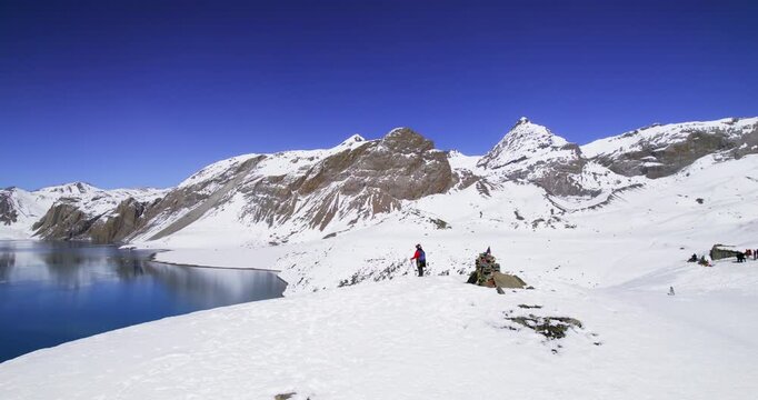 Trekkers gather at the edge of a turquoise high-altitude lake surrounded by snow-covered peaks and prayer flags in the Nepalese Himalayas.