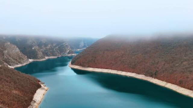 Aerial view of mountain reservoir making a bend around a sandy headland. Turquoise water bends in a shape around a headland as exposed sandy shoreline traces the full curve below foggy hills.