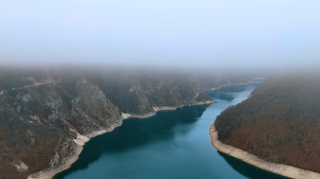 Aerial view of narrow mountain reservoir in deep rocky canyon under dense fog. The reservoir narrows into a deep canyon where limestone walls rise on both sides and dense fog fills the air above.