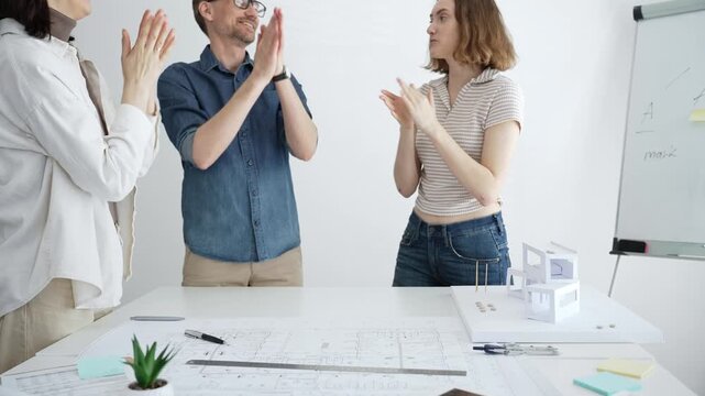 Architecture team collaborating on a project, joining hands as a symbol of unity, teamwork, and success, working on blueprints and a building model in the studio