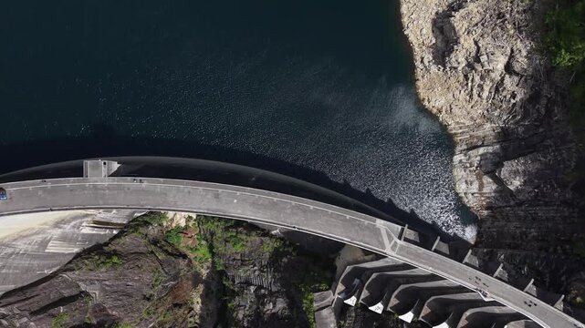 Aerial view of the massive concrete Verzasca Dam wall bordering deep blue reservoir water and steep rocky cliffs Gordola, Ticino, Switzerland.