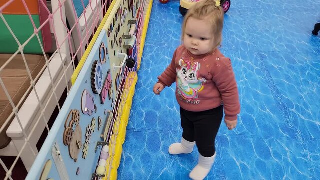 Toddler girl Sofia standing on a blue floor near a busy board wall