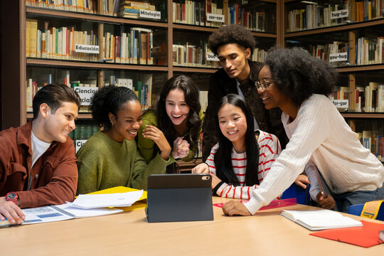 Group of diverse high school students studying and smiling around a tablet at a library table