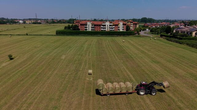 Agricultural Harvest in Summer: A tractor transports freshly baled hay across a vast, sun-drenched field, with residential buildings visible in the distance.