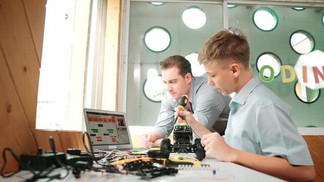 Caucasian teacher pointing at laptop screen and giving advise for programming system or coding prompt while smart high school boy listen and use equipment to repair robot at STEM classroom