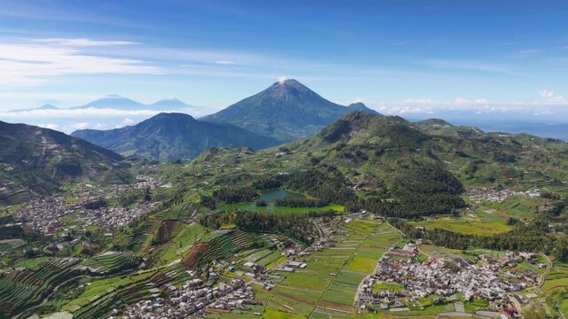 Aerial view of Dieng Plateau featuring terraced agricultural fields and the majestic Mount Sindoro under a clear blue sky in Kejajar, Central Java, Indonesia.