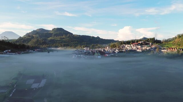 Aerial view of Dieng Plateau village nestled in a valley shrouded in thick morning mist and surrounded by rolling green hills under a bright sky Kejajar, Central Java, Indonesia.