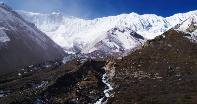 A stunning aerial view reveals towering snow-covered Himalayan peaks with a small glacial stream winding through a rugged valley under a clear blue sky.