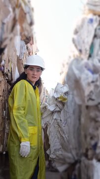 Asian Supervisor In Yellow Coat Monitoring Towering Cardboard Bales At Recycling Center, Wearing Helmet And Safety Goggles, Inspecting Baling Process, Optimistic Expression, Circular Economy Focus,