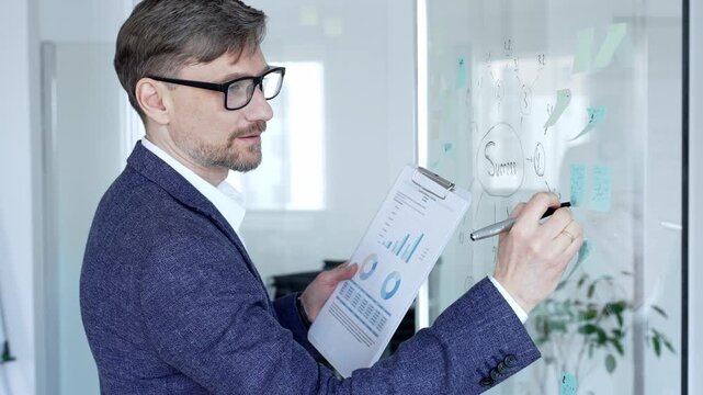 Business man evaluating success strategy roadmap on a glass wall, analyzing project development and financial data charts on a clipboard in a modern office