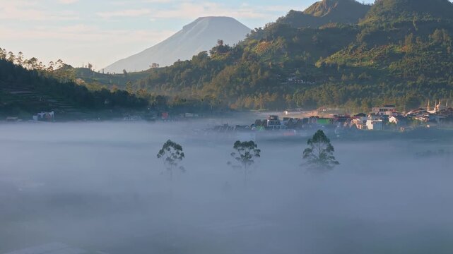 Aerial view of the Dieng Plateau village shrouded in thick morning mist with mountains and lush green hills in the background Kejajar, Central Java, Indonesia.